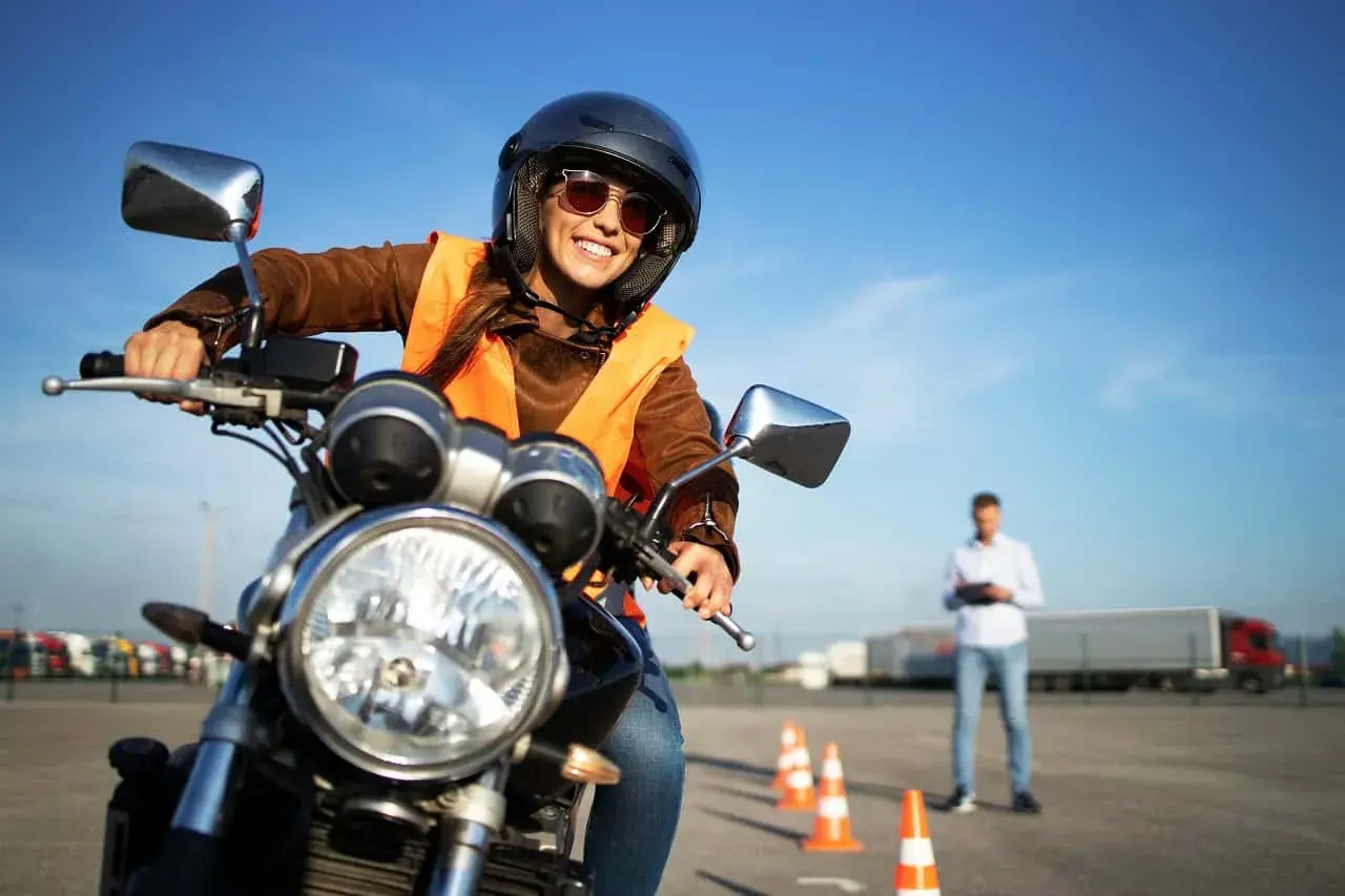 Uśmiechnięta kursantka w kasku i kamizelce na motocyklu, gotowa do jazdy. Instruktor obserwuje ją na placu manewrowym. kategoria a.