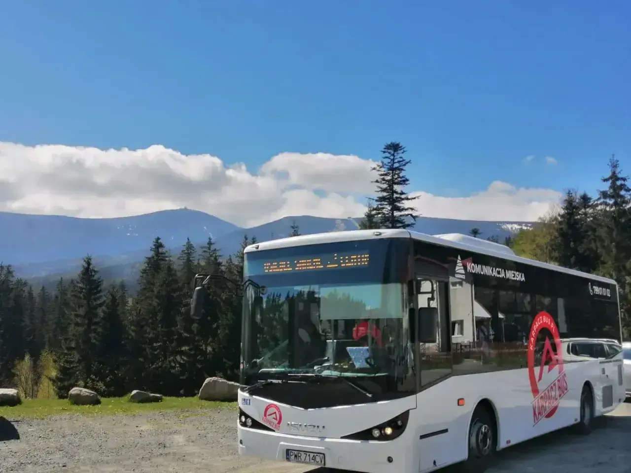 Biały autobus "Karpacz Bus" z napisem "Komunikacja Miejska" na tle gór i lasu. Zastanawiasz się, ile kosztuje autobus?