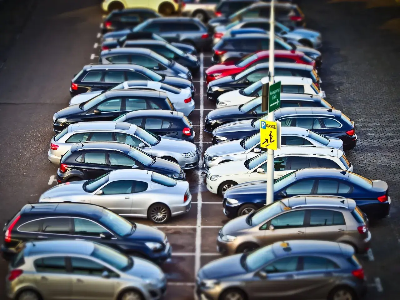 Rząd samochodów na parkingu. Znak "Kasse" sugeruje, że to parking płatny. KNF co to? To może być skrót od nazwy firmy zarządzającej parkingiem.