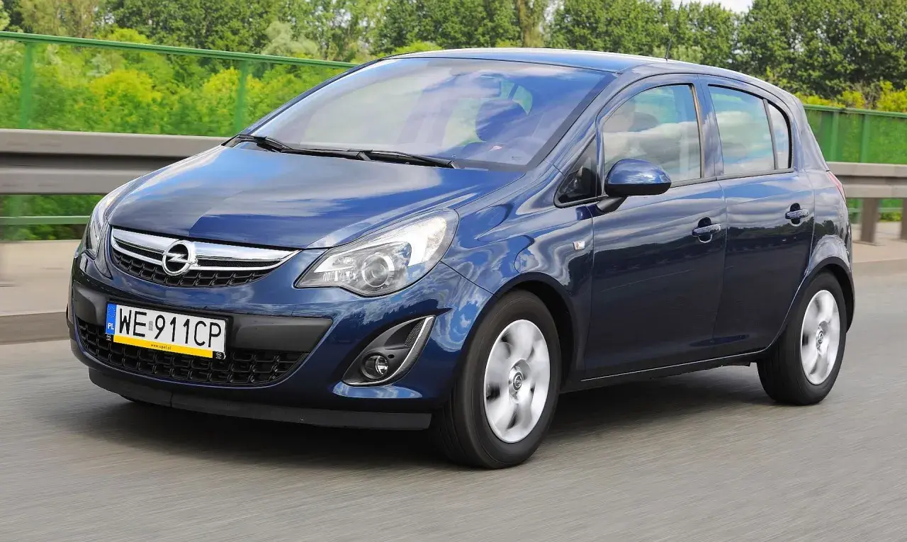 Niebieski Opel Corsa D 1.4 benzyna w ruchu drogowym, z zielenią drzew w tle.
