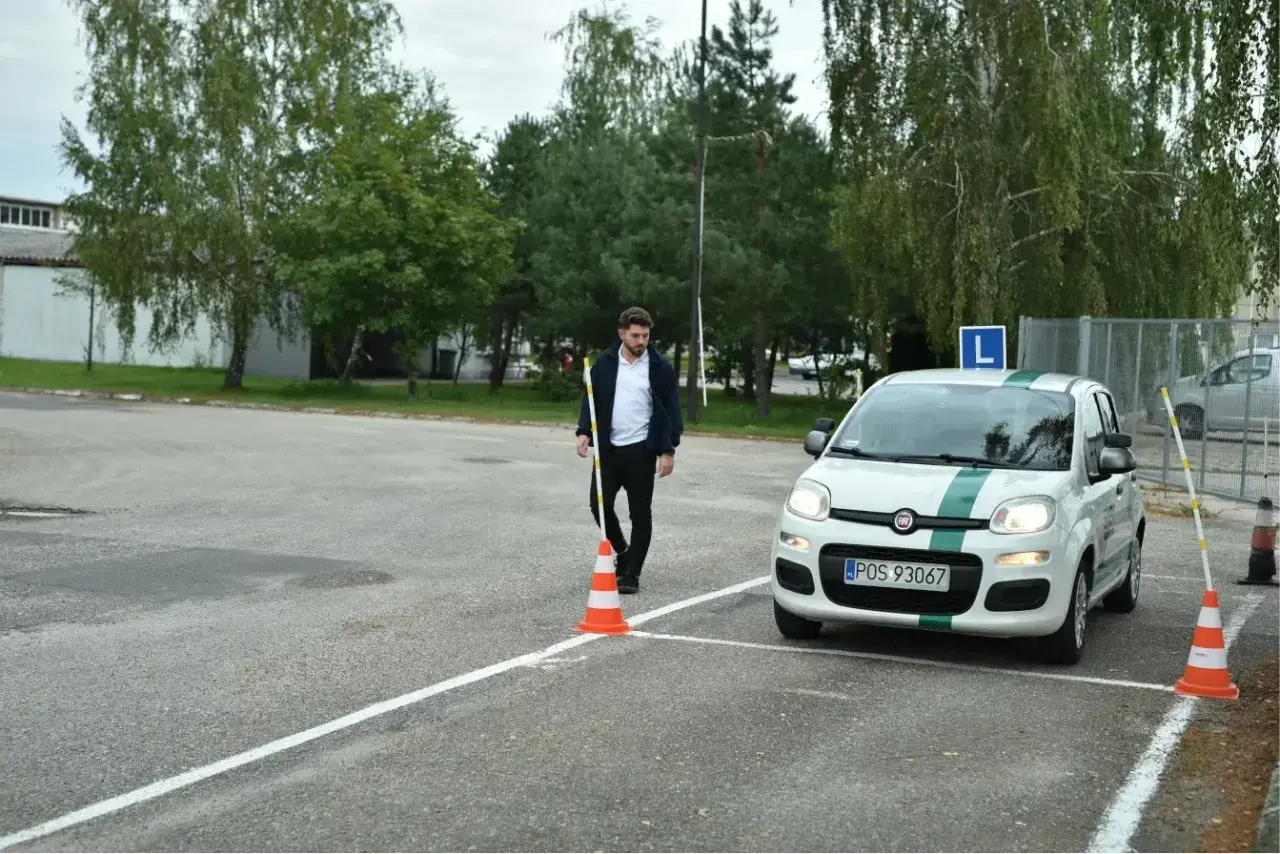 Młody mężczyzna ustawia pachołek na placu manewrowym. Obok stoi biały Fiat Panda z literą "L" na dachu, gotowy do egzaminu.
