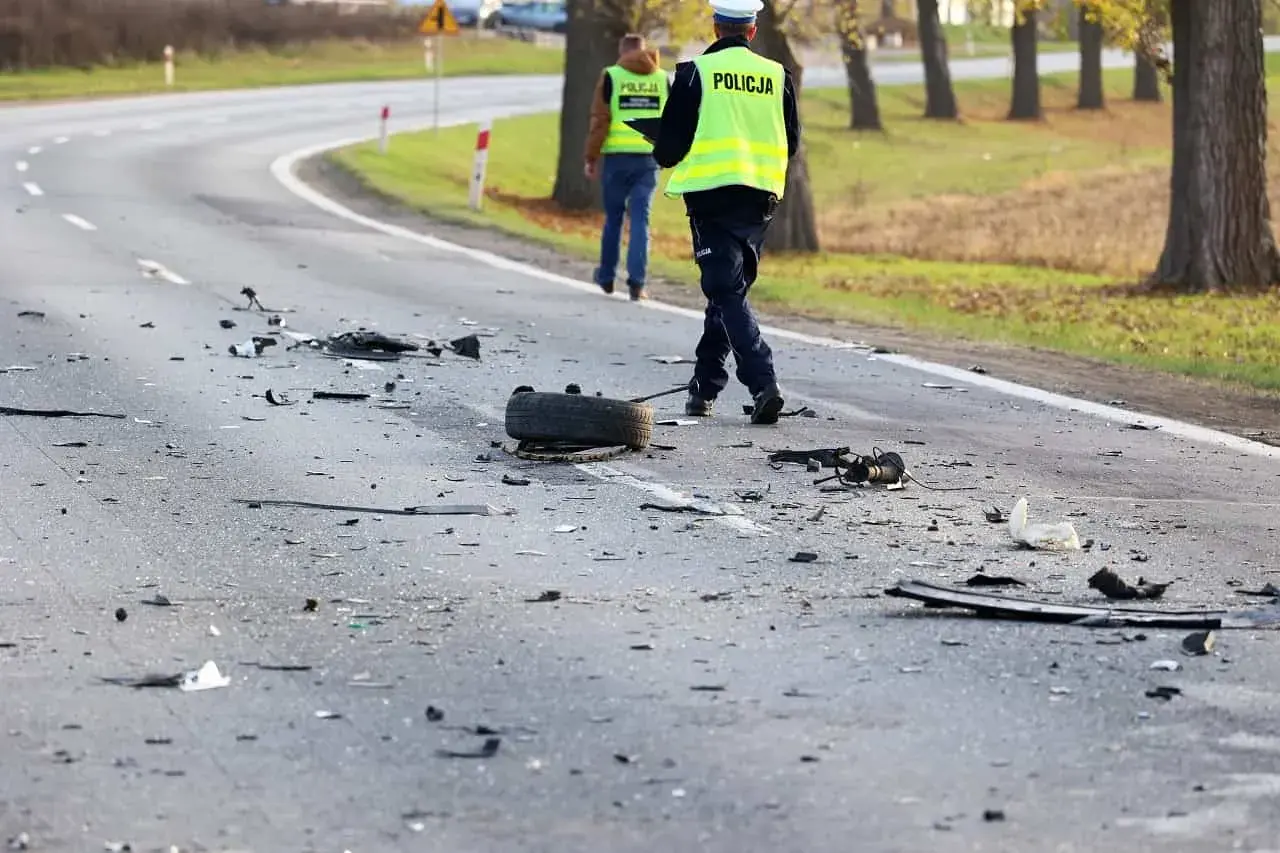 Na drodze po kolizji leżą szczątki pojazdów i opona. Policjanci w kamizelkach odblaskowych badają miejsce zdarzenia, być może szukając śladów ucieczki z miejsca kolizji.