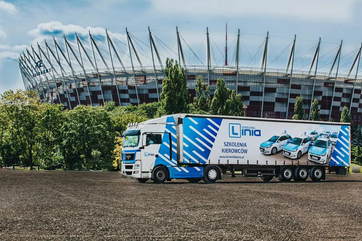 Ciężarówka z logo "Linia Szkolenia Kierowców" i samochodami na naczepie, na tle Stadionu Narodowego. Marzysz o **prawie jazdy na tira**? Tu możesz je zdobyć!
