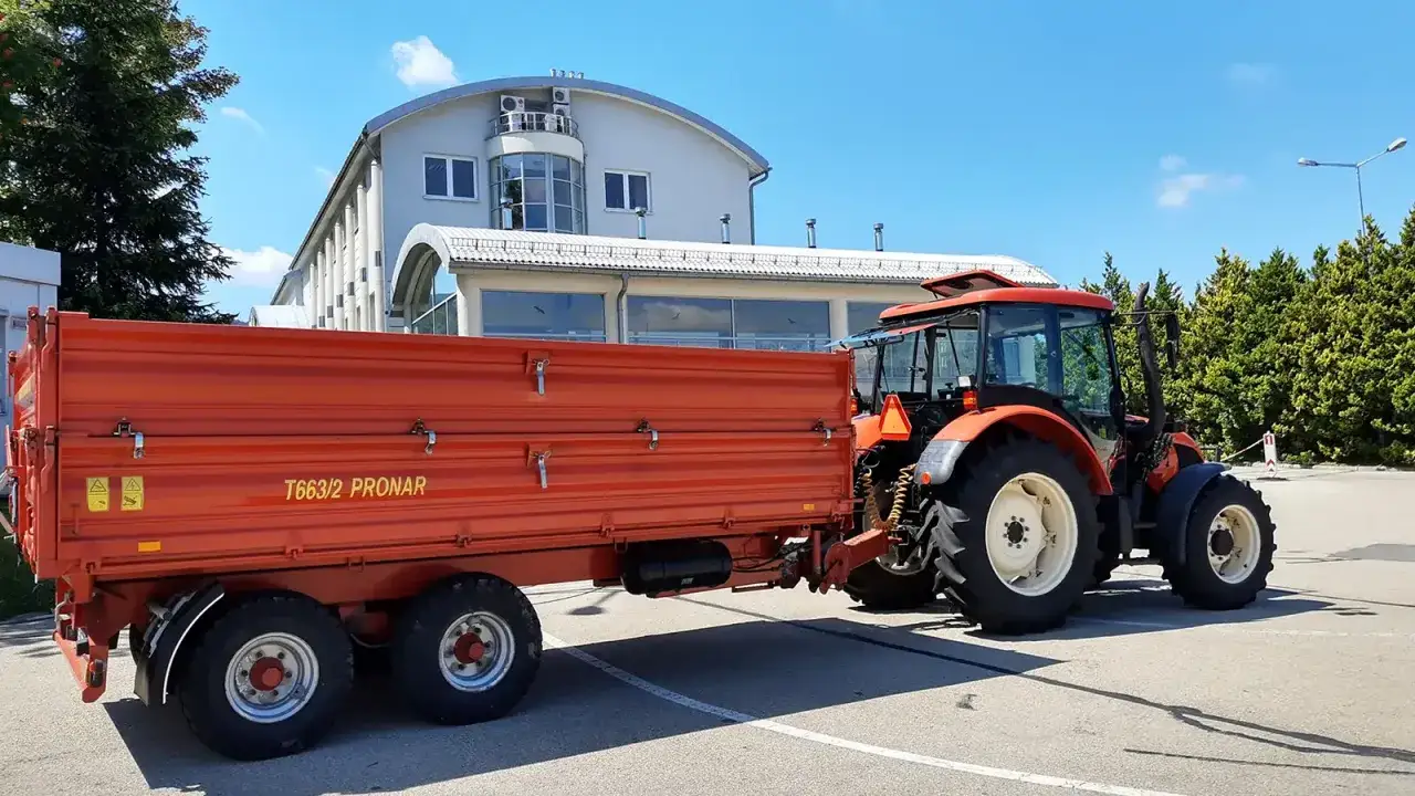 Pomarańczowy traktor z przyczepą Pronar T663/2, gotowy do pracy. Posiadanie prawa jazdy kat T to klucz do obsługi takiego sprzętu.
