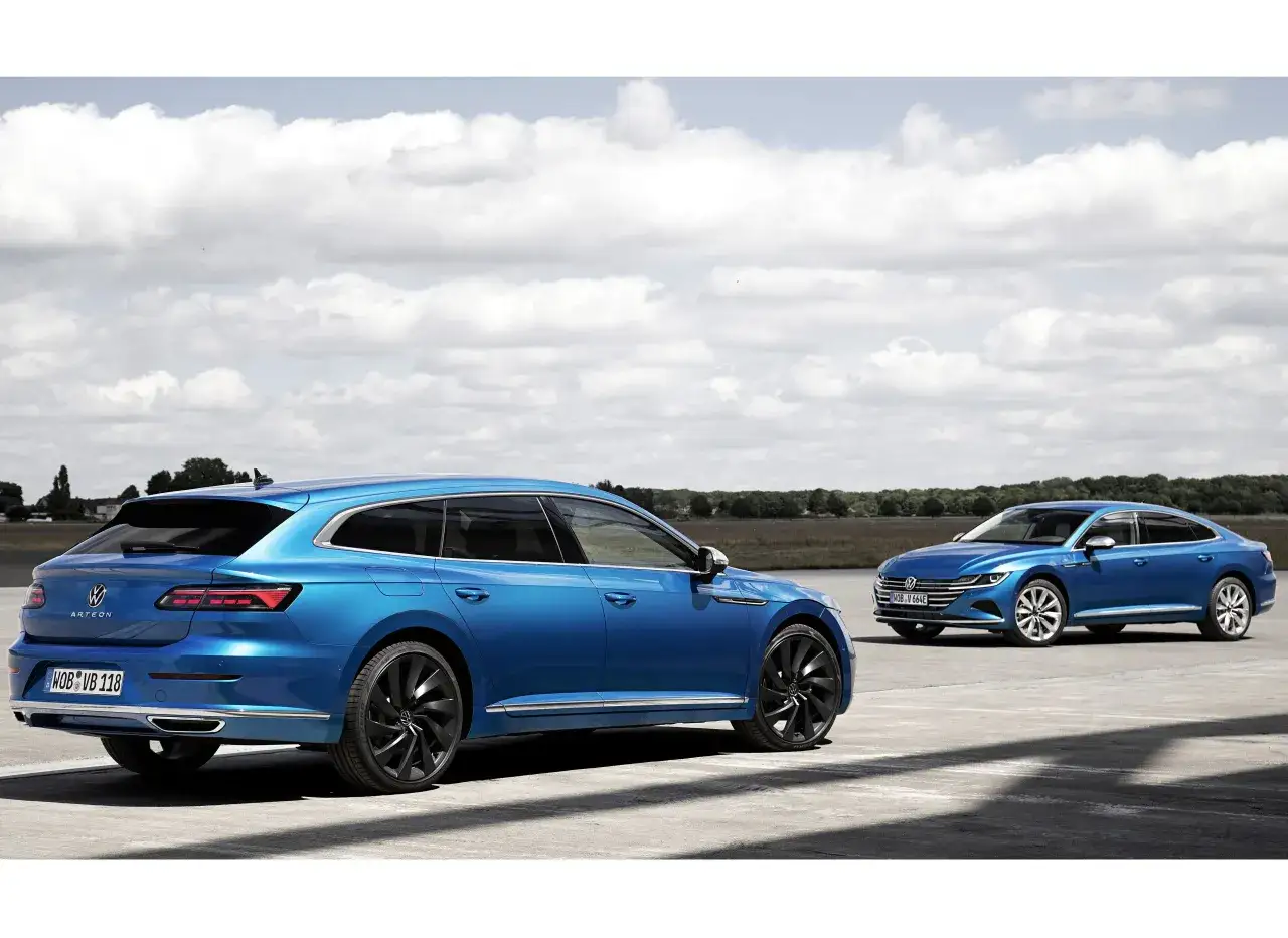 Dwa niebieskie Volkswageny Arteon Shooting Brake stoją na betonowym placu pod błękitnym niebem z chmurami.